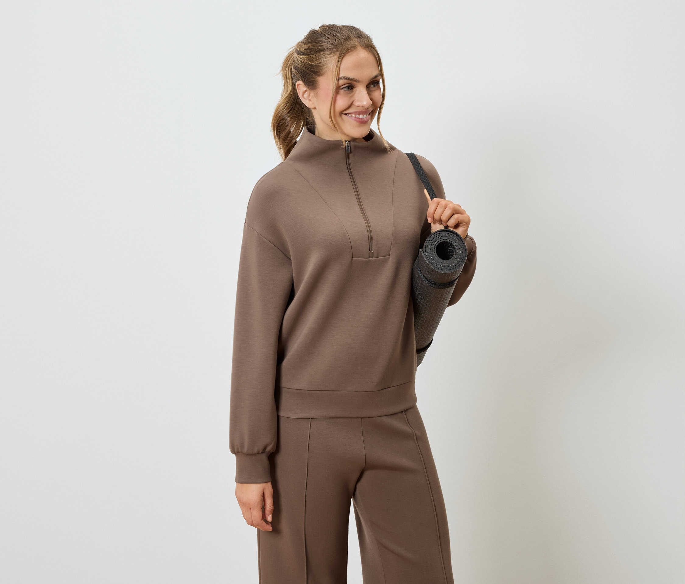 Nő barna tréningruhában, jógamatraccal a vállán.