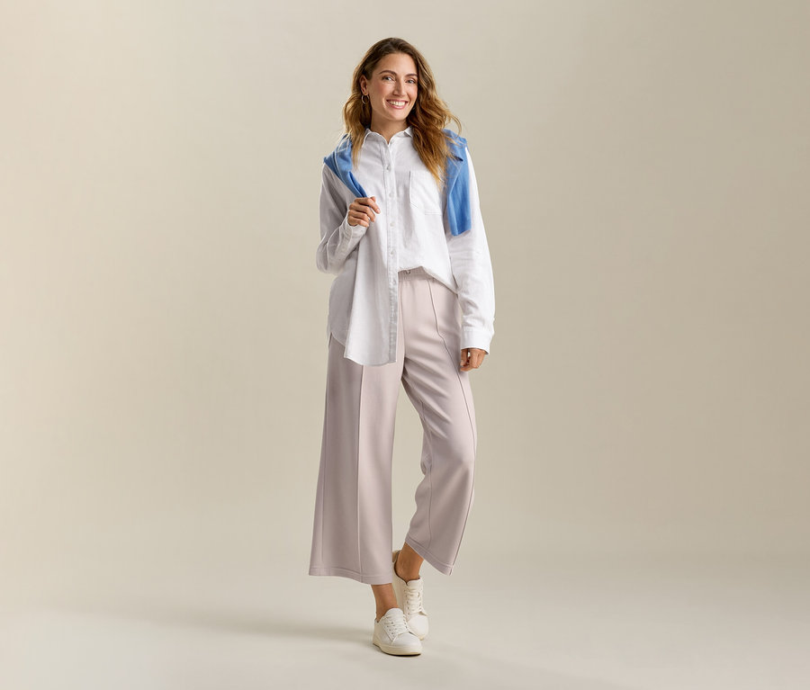 Mosolygó nő fehér, hosszú vászonblúzt és homokszínű, modálkeverékből készült, bő szárú nadrágot visel. A vállán kék pulóver van.