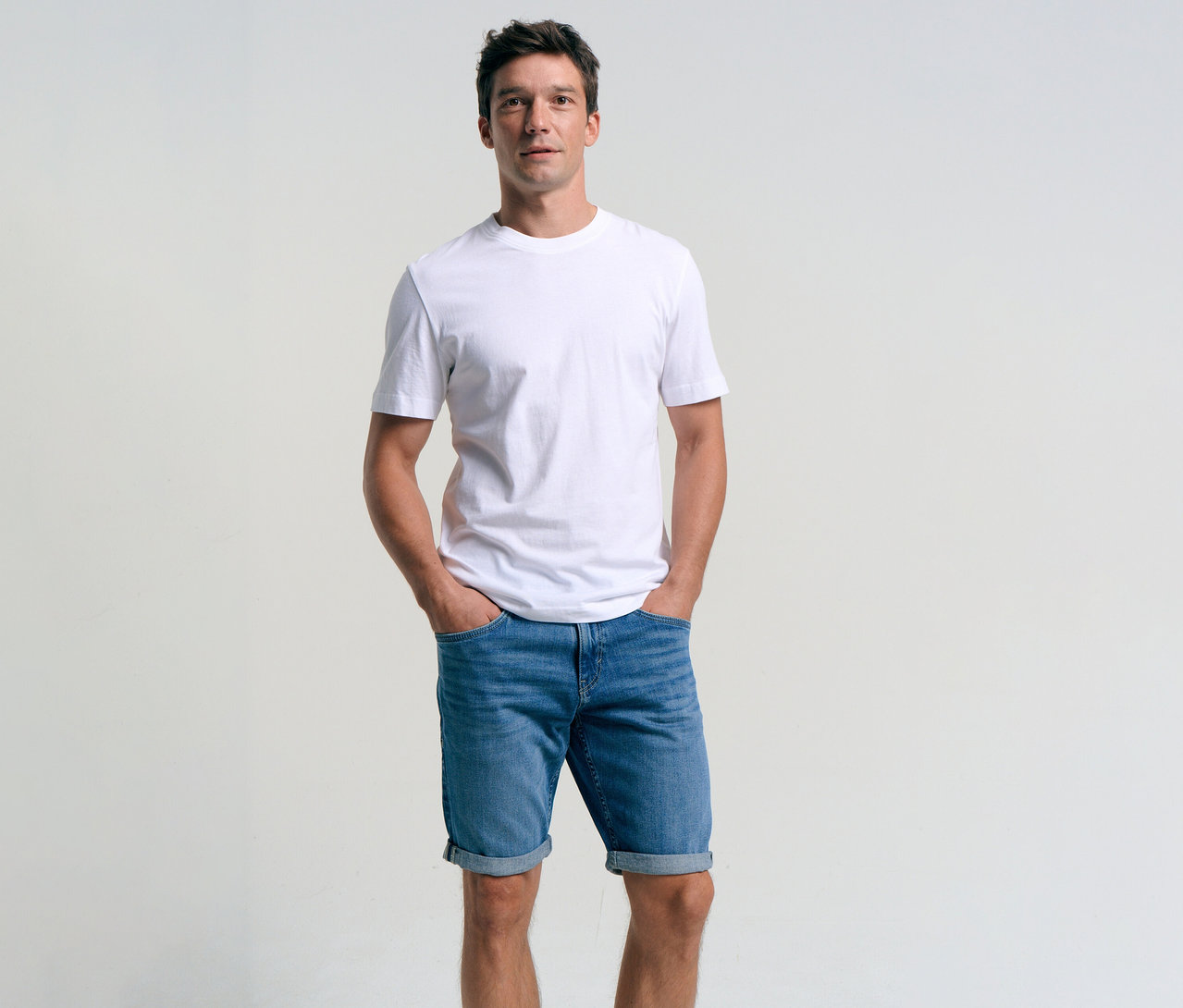 Férfi fehér pólóban és Tom Tailor TTJOSH REGULAR SLIM kőmosott középkék farmer rövidnadrágban, keze a zsebében.