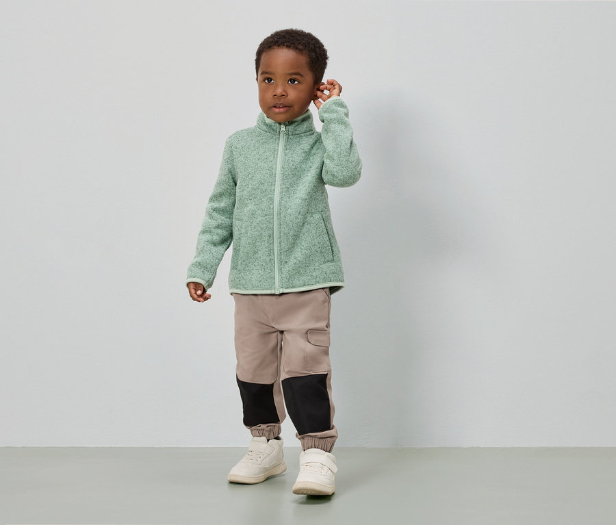 Egy fiú áll egy fehér fal előtt, világoszöld fleece kabátot, bézs színű, fekete betétekkel ellátott nadrágot és fehér cipőt visel.