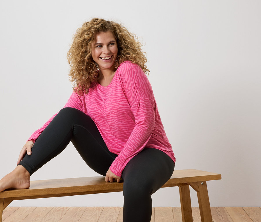 A göndör hajú nő mezítláb ül egy fa padon, rózsaszín, zebracsíkos hosszú ujjú inget és fekete leggingset visel.