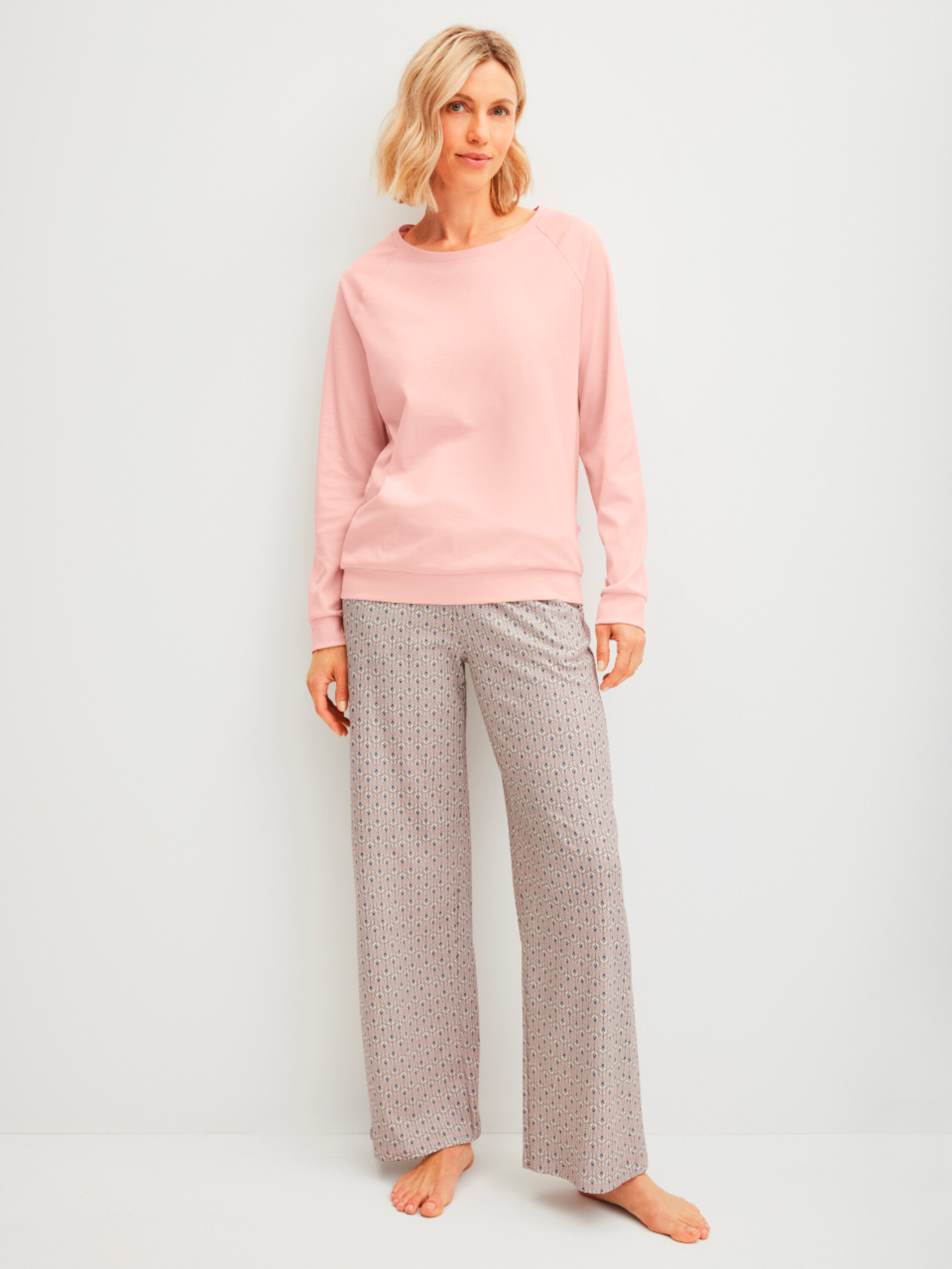 Nő áll mezítláb rózsaszín pulóverben és mintás nadrágban egy fehér fal előtt.