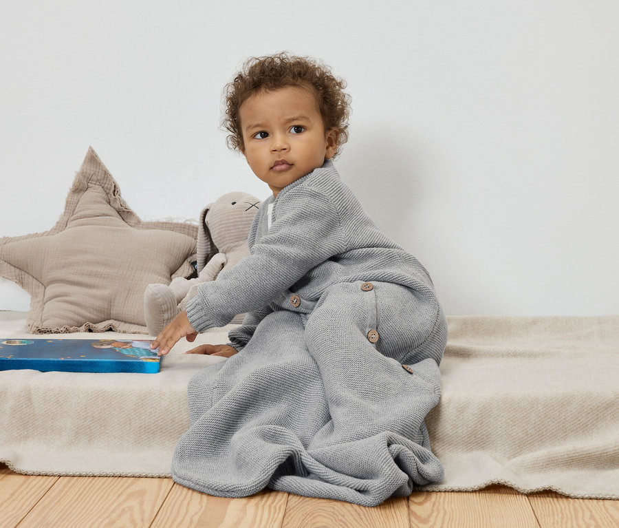 A baba egy bézs takarón ül egy csillag és egy nyuszi plüssállat mellett, és egy kék könyvet érint.