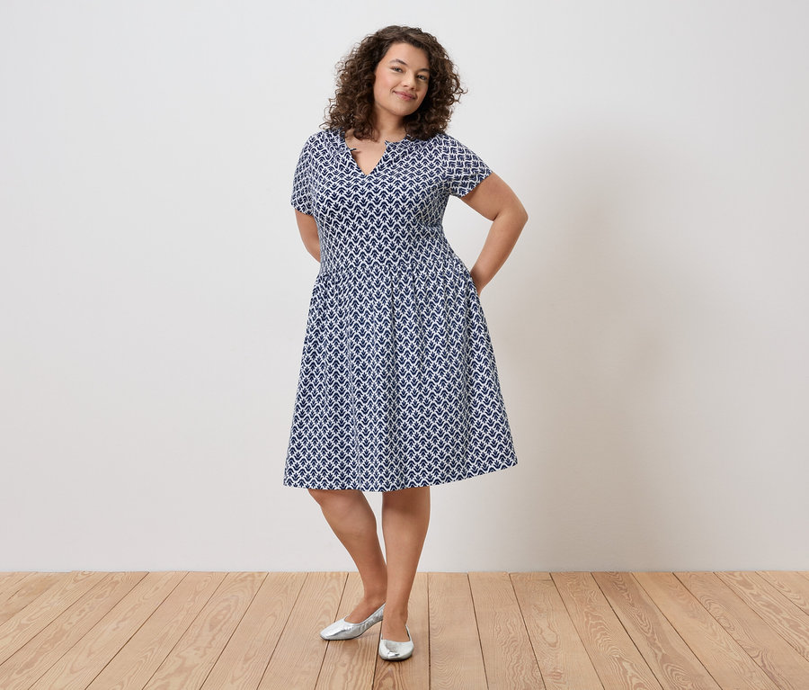 Göndör hajú nő kék-fehér ruhában és ezüst cipőben áll a fal előtt, kezével a zsebében.