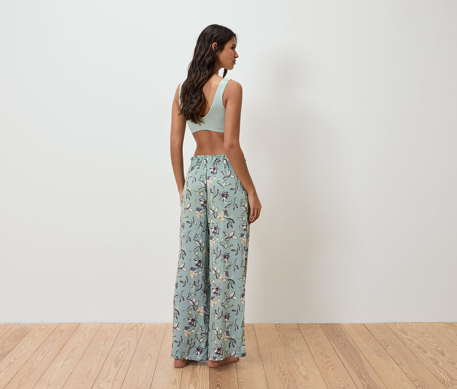 Egy nő áll háttal a kamerának, világoszöld felsőt és virágmintás, bő nadrágot visel.