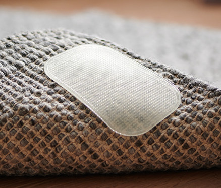 Egy szürke szőnyeg sarkának közeli képe, melyen egy áttetsző 4 csúszásgátló szőnyeghez látható.