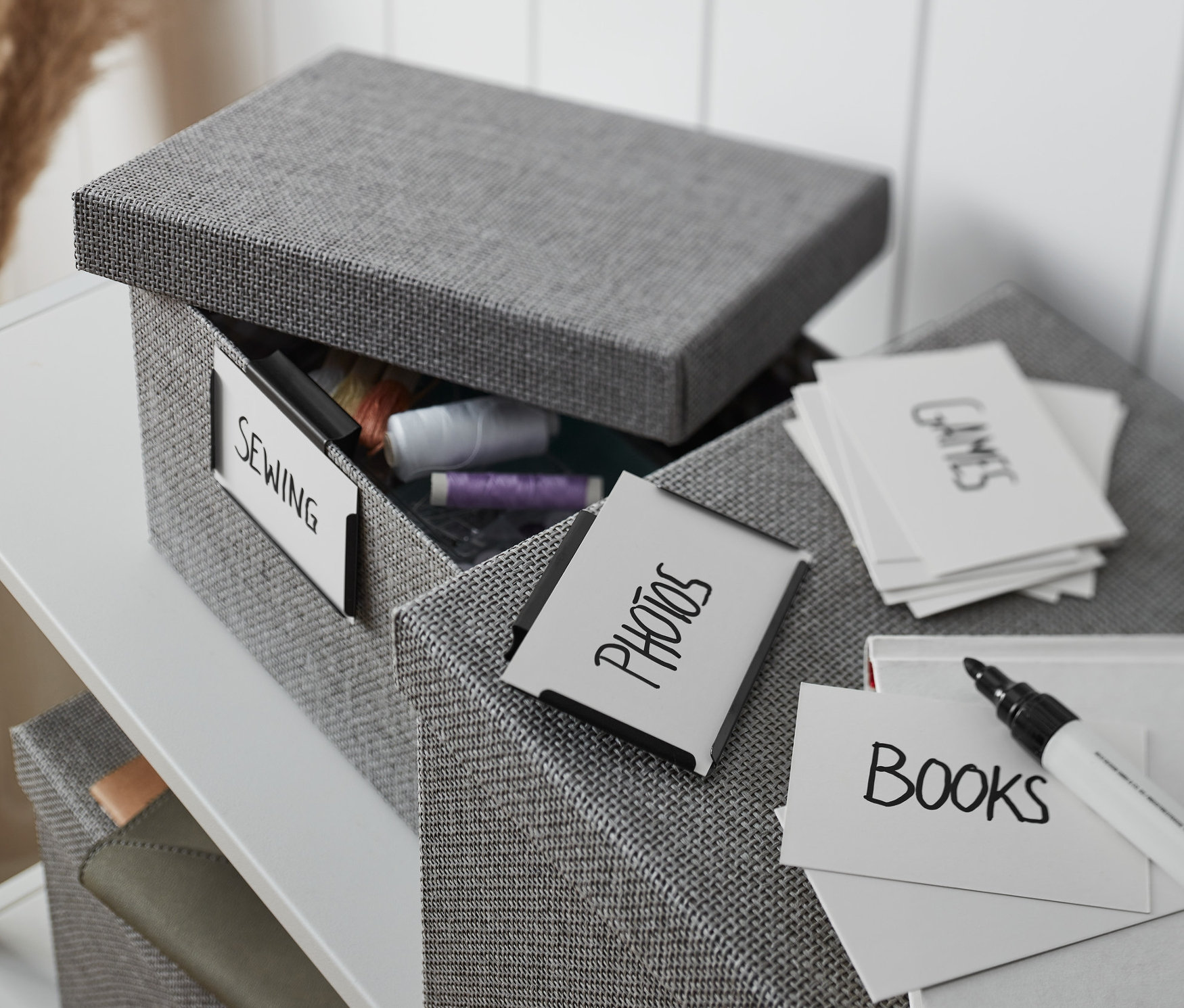 Nyitott szürke szövetdoboz 2 fém címketartóval, "varrás" felirattal és fonal tartalommal, mellette feliratozott kártyákkal ellátott doboz.
