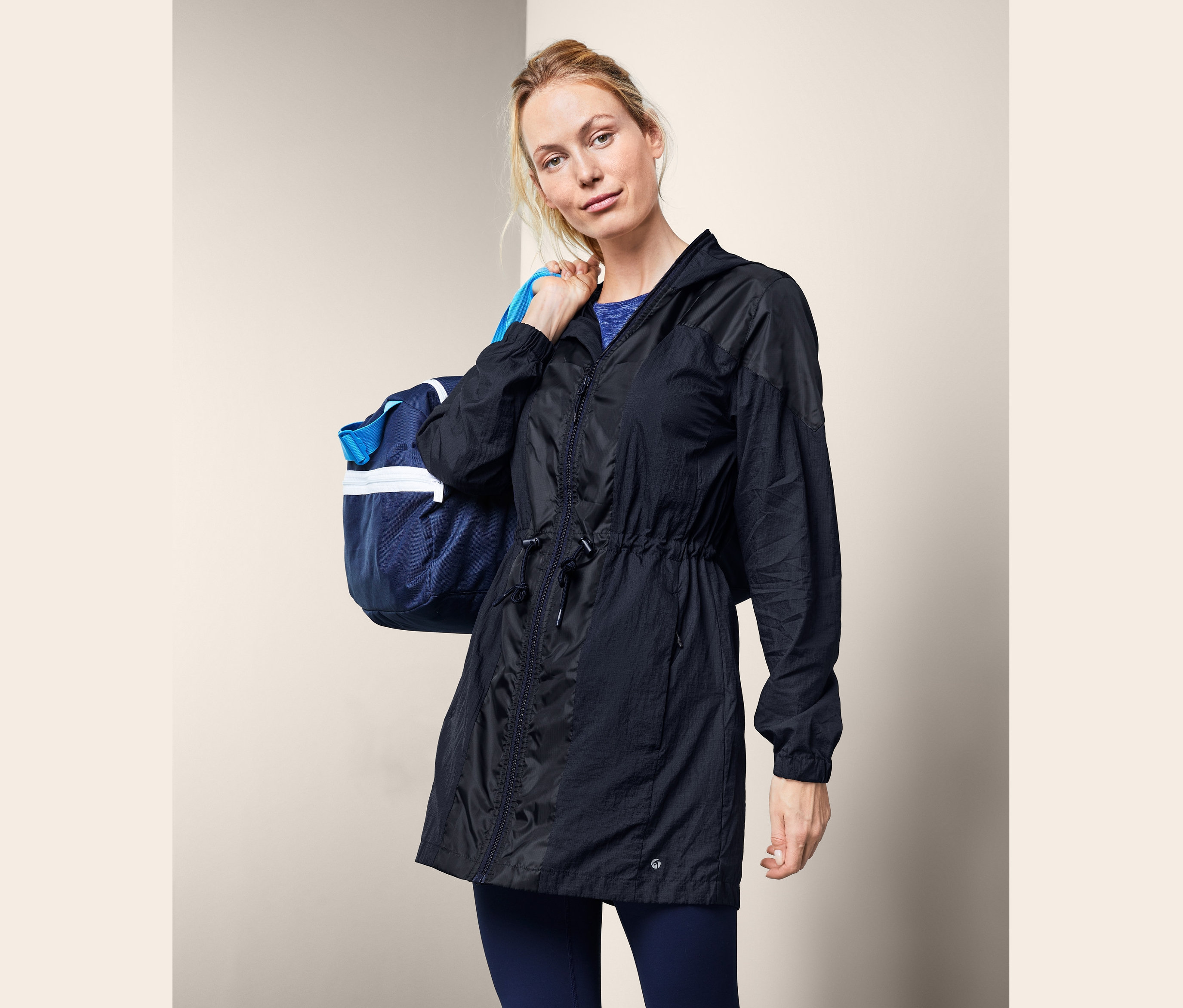 A szőke nő sporttáskát visz a vállán, és sötétkék funkcionális kabátot visel.