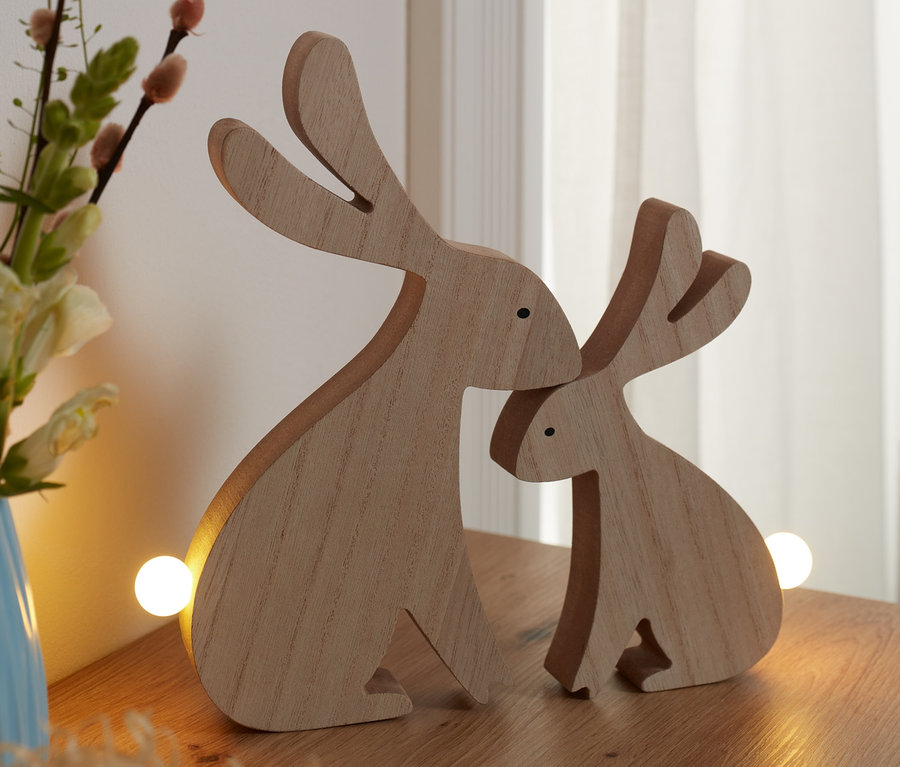 Két LED-es dísznyuszi áll egy fa asztalon egy virágvázával és egy tojáskosárral szomszédságában.