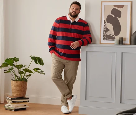 Férfi a falnak támaszkodva fehér férfi sneaker cipőt, barna férfi chino nadrágot és csíkos, sötétkék/piros férfi hosszú ujjú pólóinget visel.