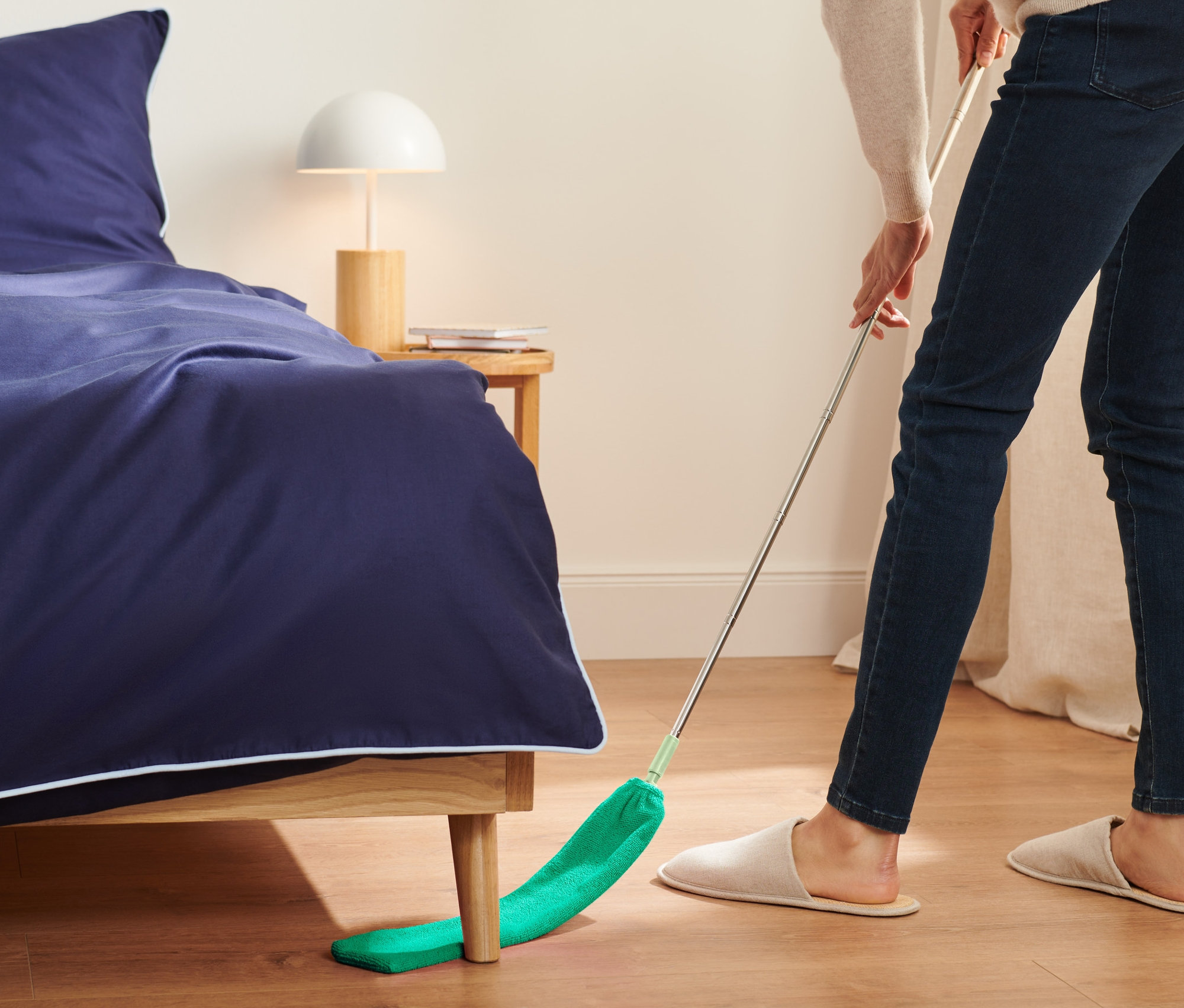 Nő a teleszkópos nyéllel ellátott portörlővel takarít az ágy alatt.