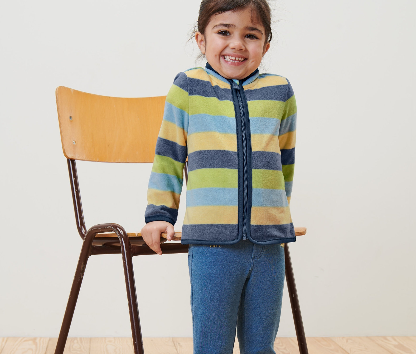 Egy mosolygós lány áll egy szék mellett és csíkos gyerek polárfelsőt visel.