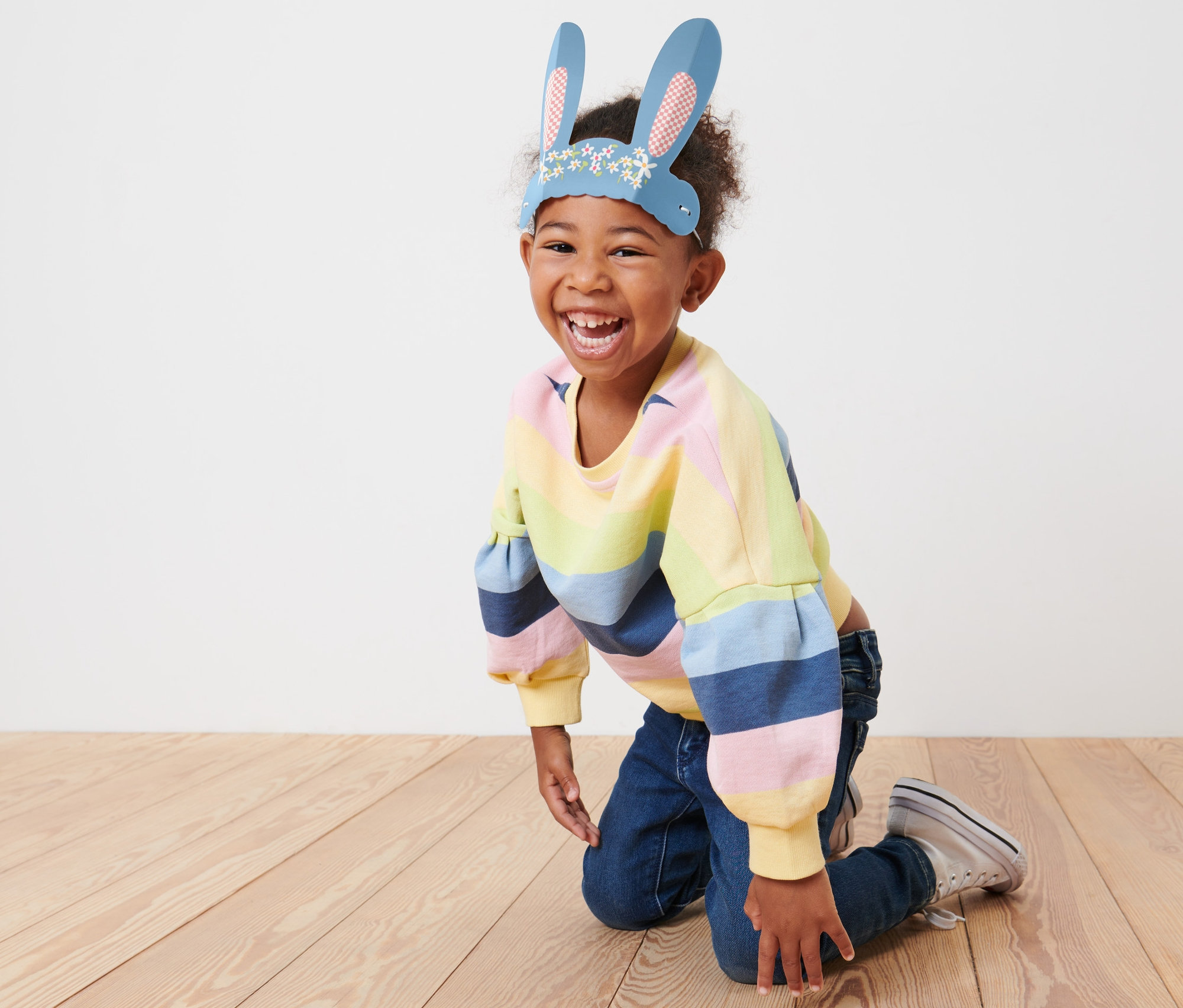 Mosolygó gyermek térdel a padlón, papír nyuszifüleket visel.