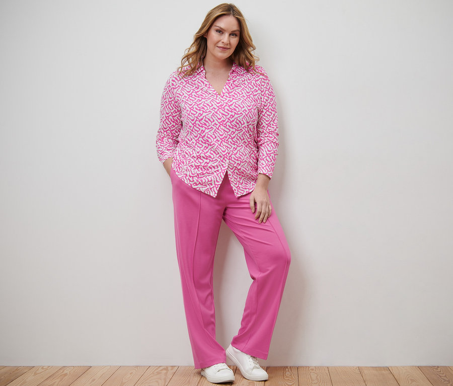 Nő pózol női pólóblúzban gomboláspánttal és jersey nadrágban levarrt hajtással.