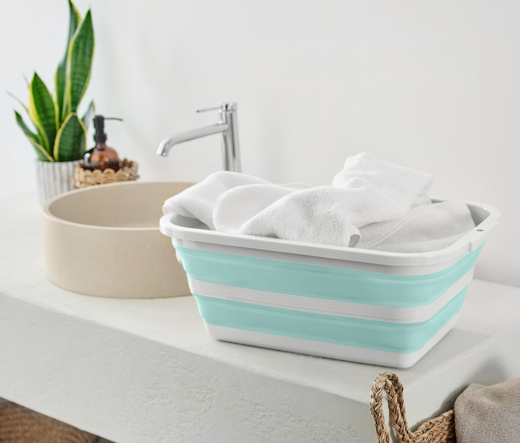 Egy összehajtható ruháskosár tele fehér törölközőkkel egy fehér polcon áll a mosdókagyló mellett.