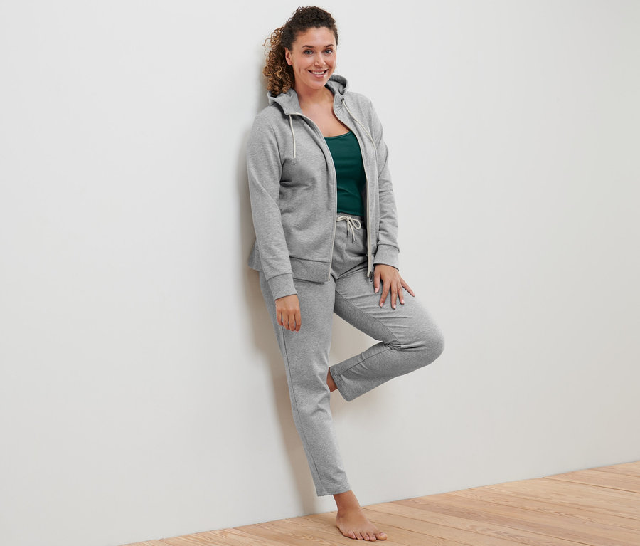 Egy nő a falnak dőlve szürke kapucnis szabadidőfelsőt, 2 zöld és törtfehér női topot és szürke női szabadidőnadrágot visel.