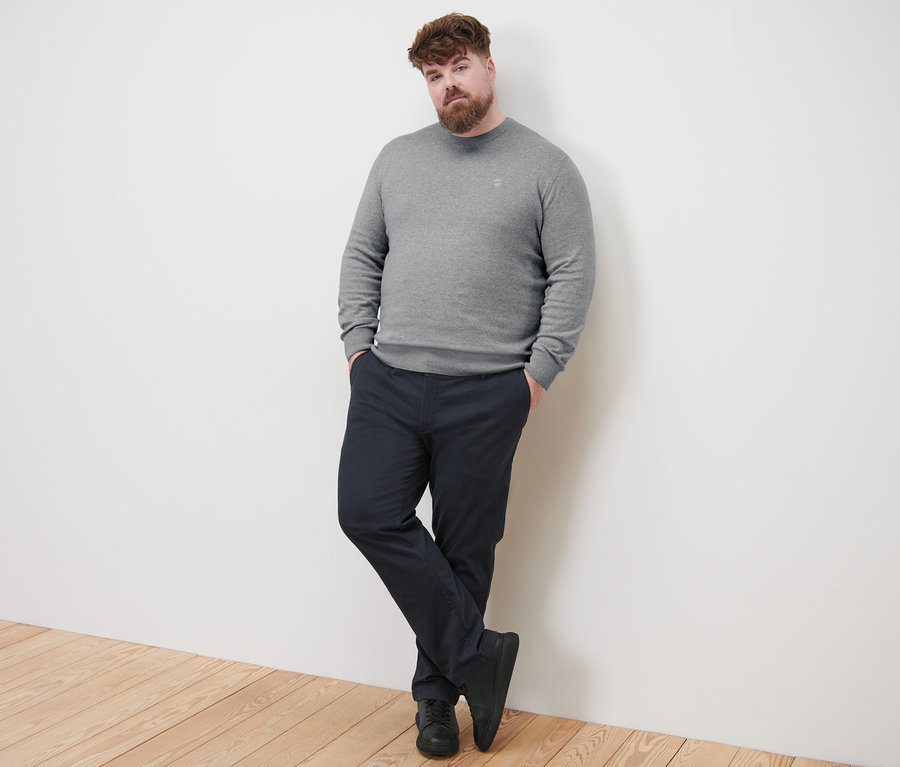 Férfi támaszkodik a falnak, szürke kötött pulóvert és tengerészkék chino nadrágot visel.
