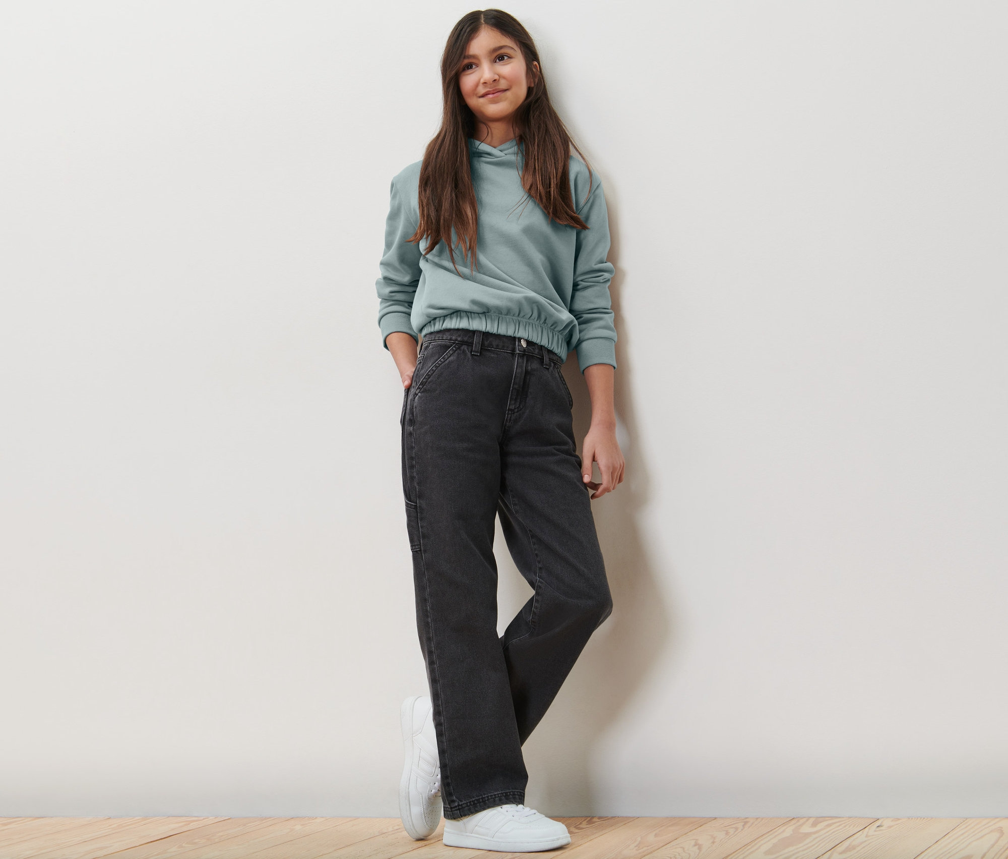 Egy lány gyerek oldalzsebes nadrágban és kapucnis pulóverben pózol.
