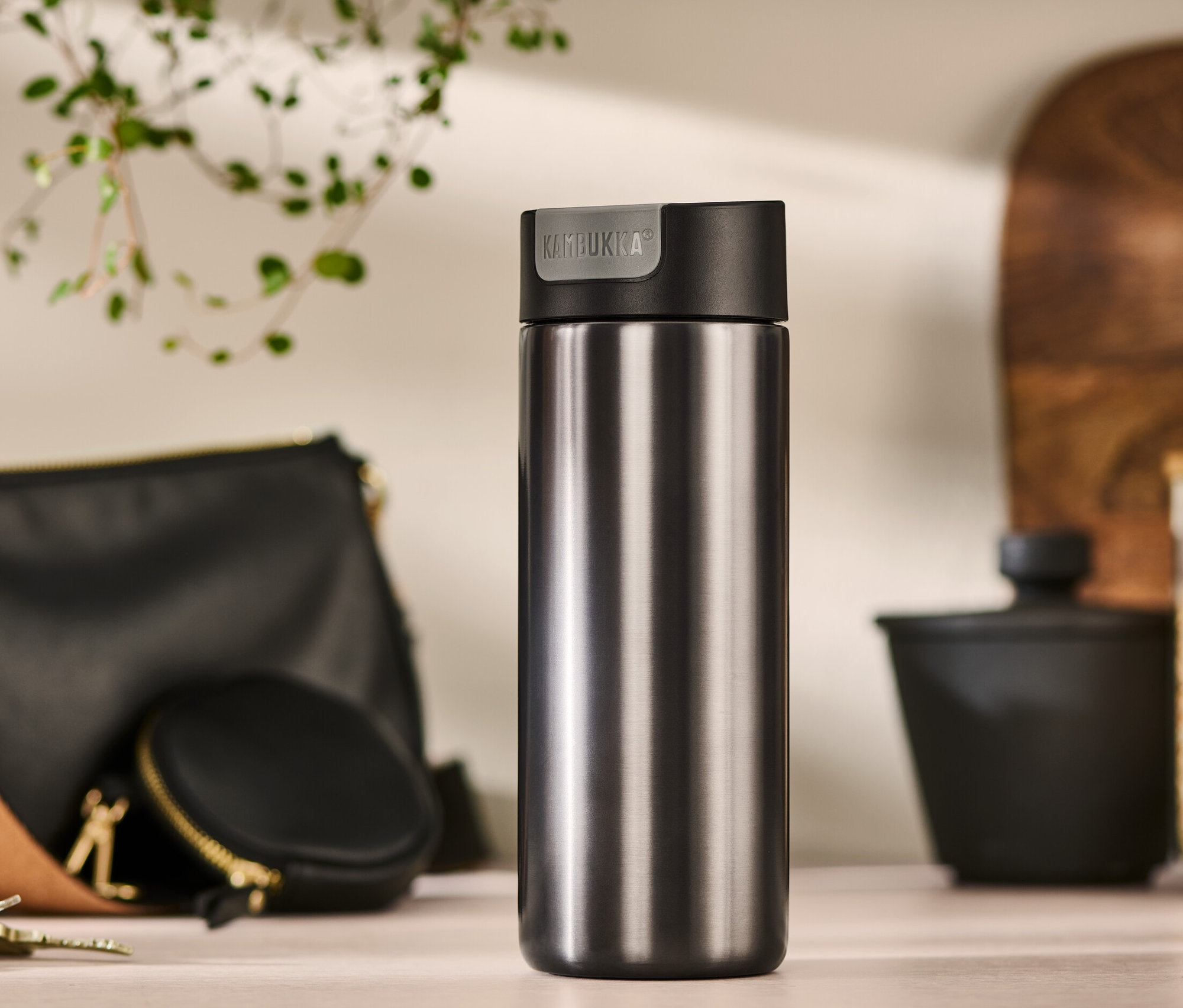 Egy szürke, 500 ml-es Kambukka To-Go bögre áll az asztalon egy kézitáska, egy pénztárca és kulcsok mellett.