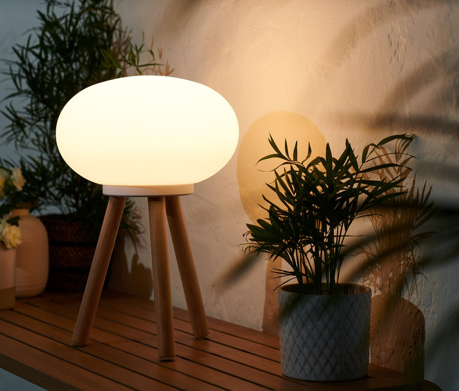 A kültéri LED-es asztali lámpa fa lábakkal egy fa asztalon áll egy cserepes növény mellett.