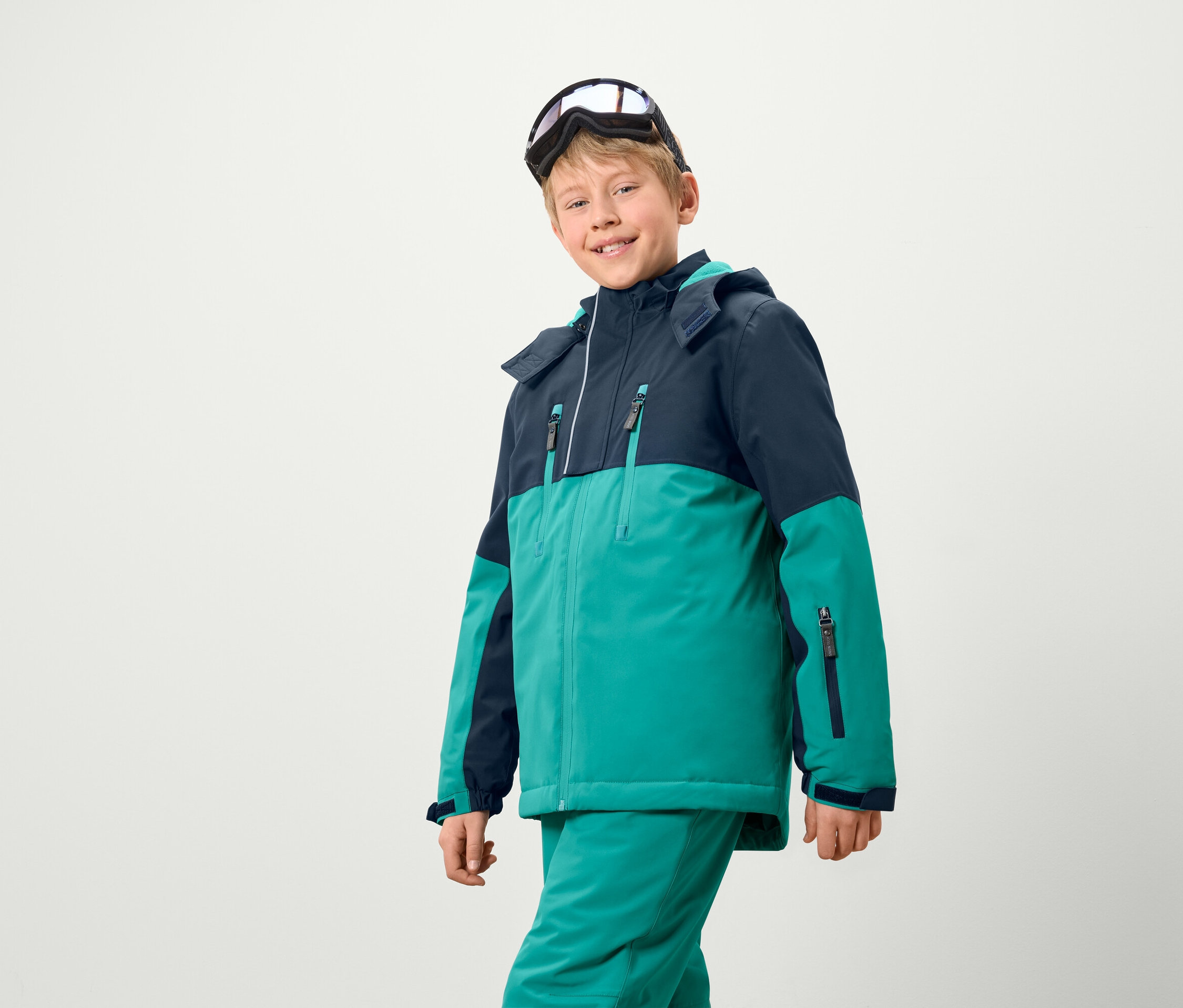 Egy fiú síkabátot és -nadrágot visel, sí szemüveggel a fején.