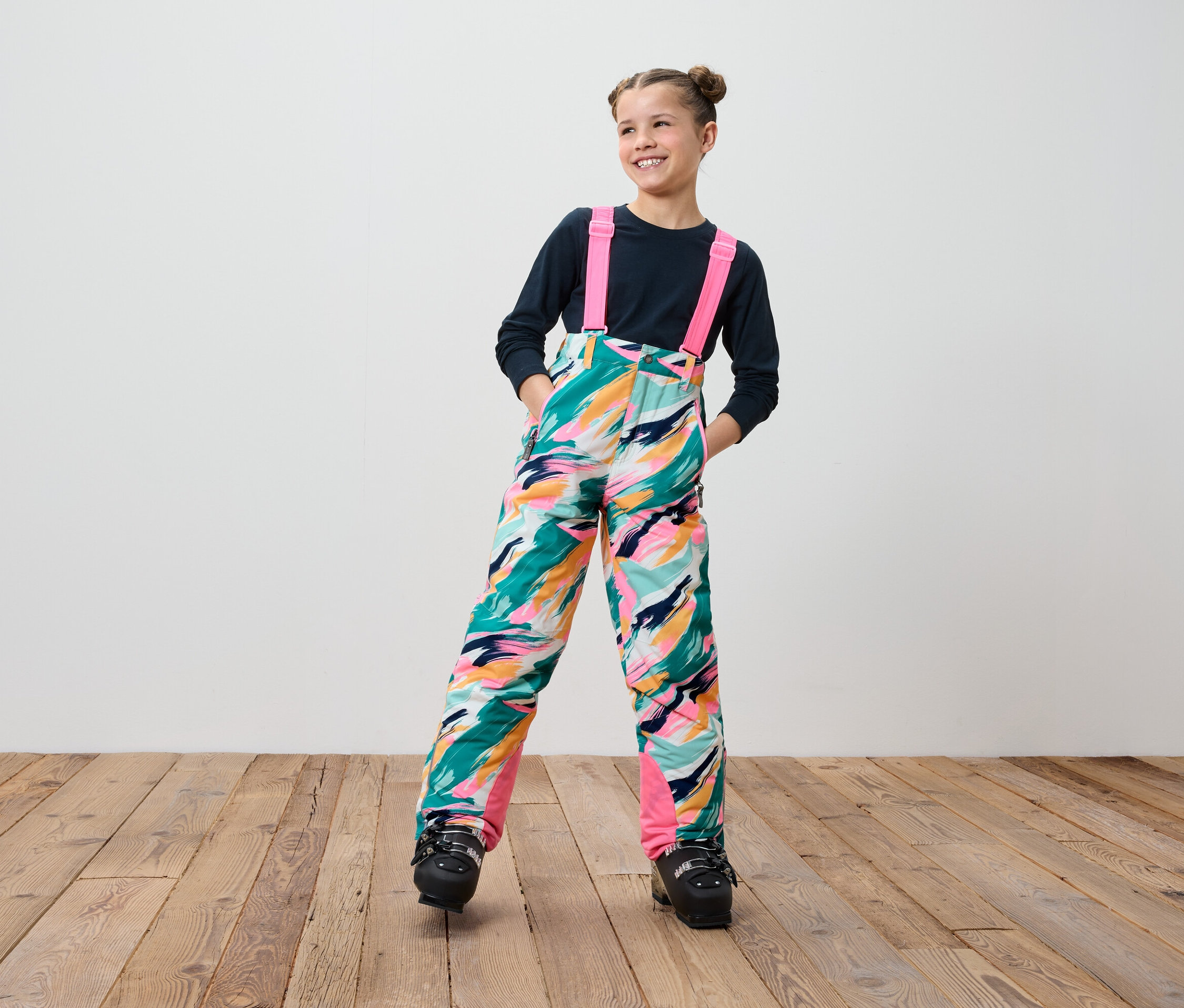 Mosolygó tinédzser lány színes kantáros sí nadrágban és fekete síbakancsban áll fa padlón.