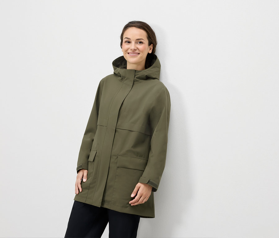 Nő pózol olívazöld kapucnis kabátban és fekete nadrágban.