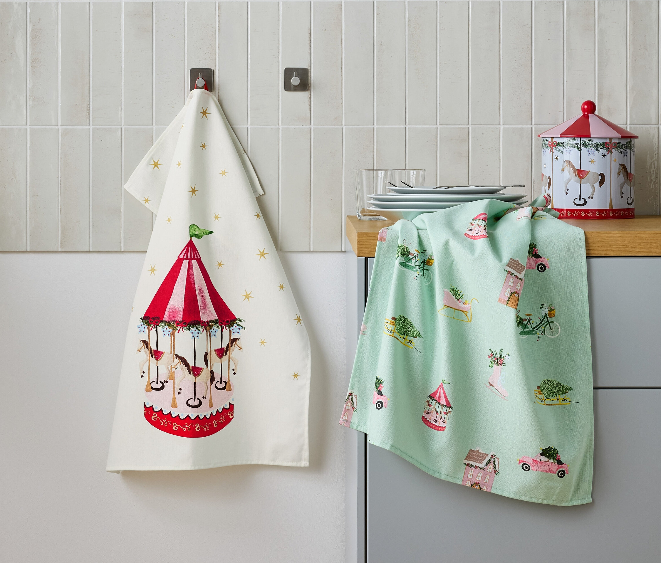 Két konyharuha lóg egymás mellett a falon, az egyik karusszel mintával, a másik ünnepi motívumokkal, valamint egy fémdoboz az asztalon.