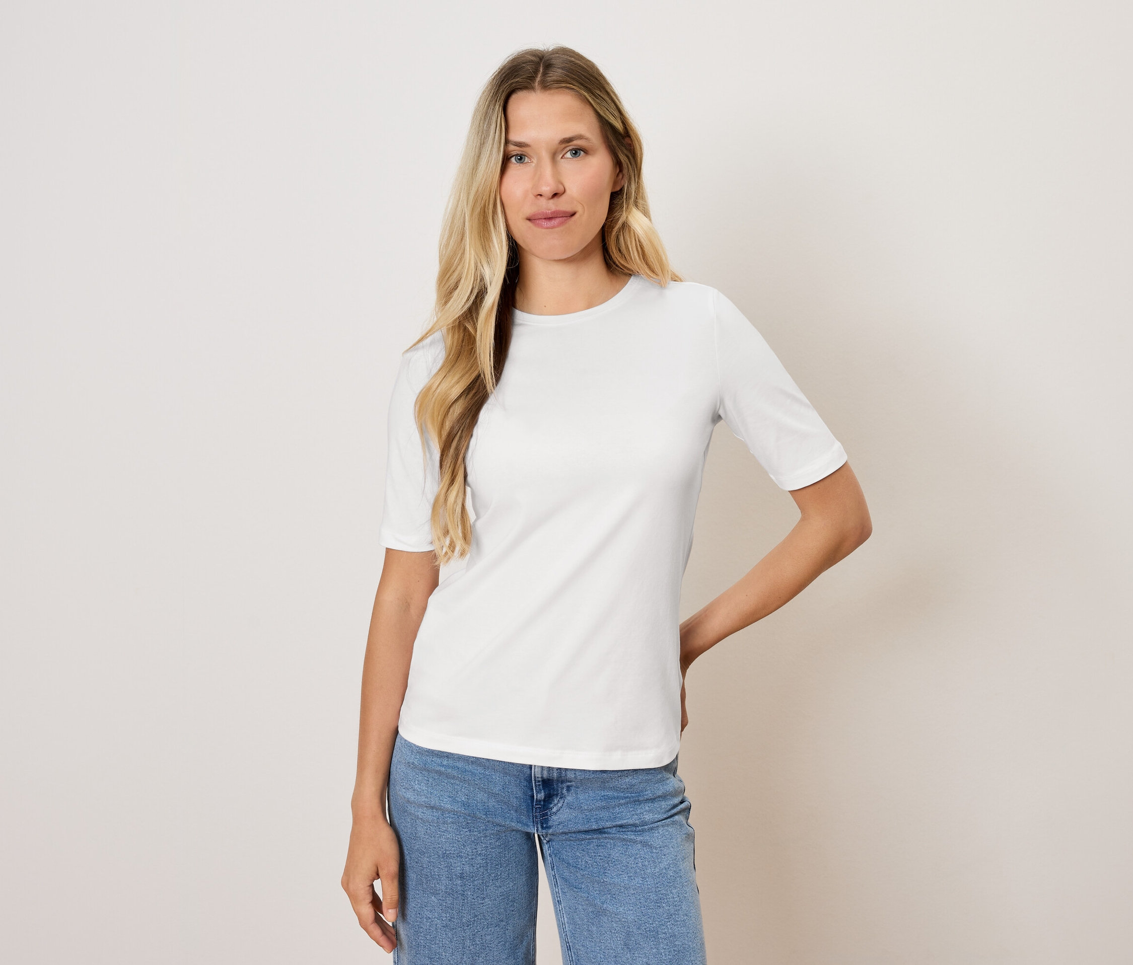 Szőke hajú nő pózol fehér pólóban és kék farmerban.