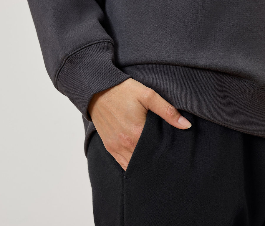 Egy nő részlete, amint a kezét a fekete szabadidőnadrágjának zsebébe teszi, szürke pulóverrel.