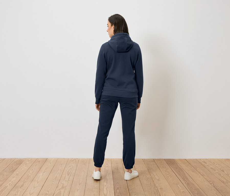 Hátulról látható nő kék kapucnis pulóverben és nadrágban, fa padlón áll egy fehér fal előtt.