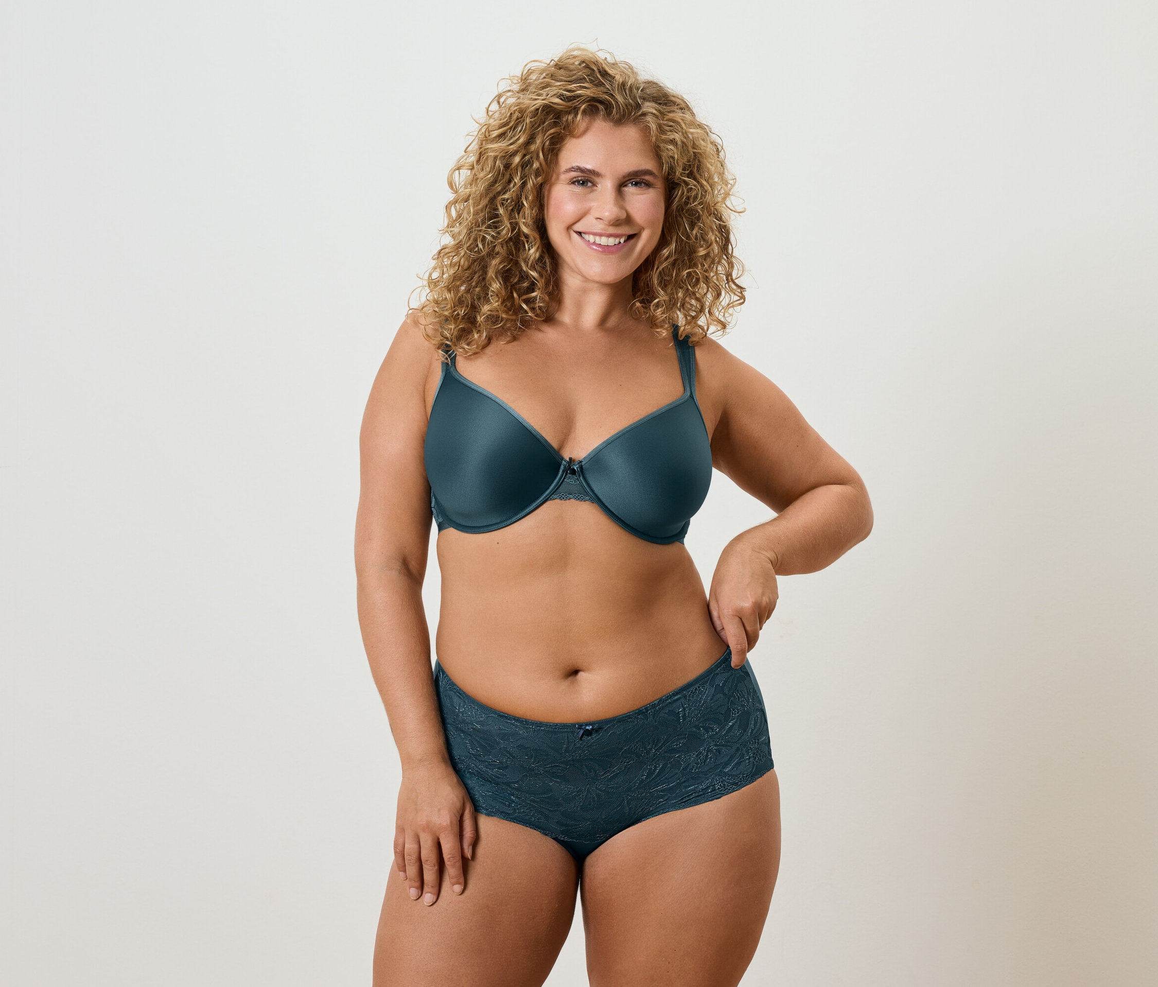 Göndör szőke hajú nő petróleum színű melltartót és hozzáillő alsóneműt visel.