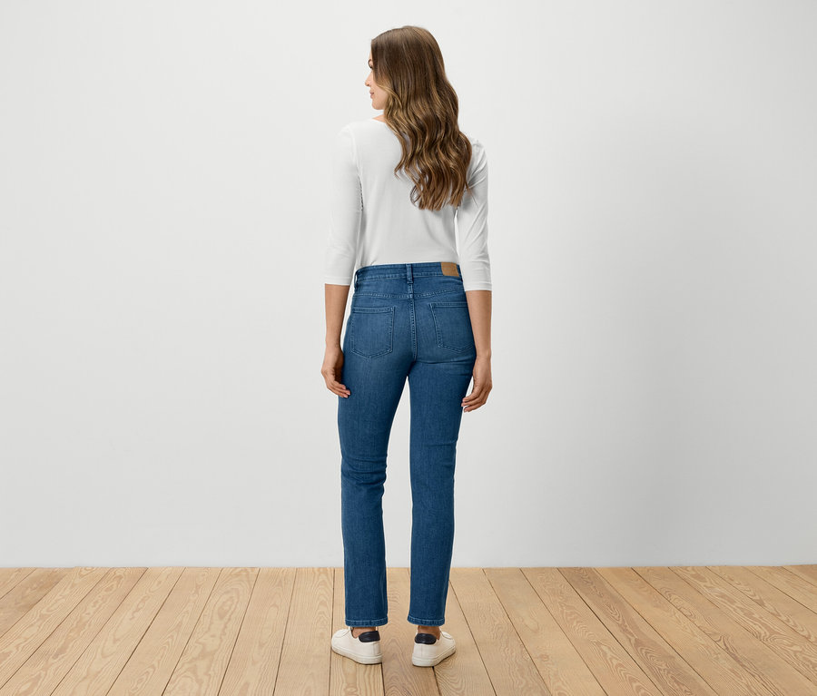 Egy nő háttal áll a kamerának, kék farmerben, fehér ingben és fehér cipőben.