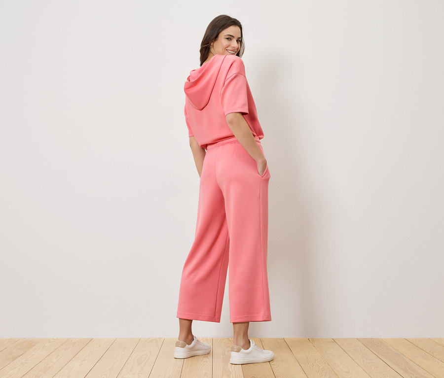 A vállán átnéző nő rózsaszín kapucnis pulóvert és culotte nadrágot visel.