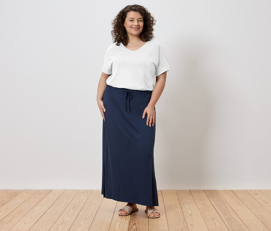 Nő áll, fehér pólót, hosszú, sötétkék szoknyát és szandált visel.