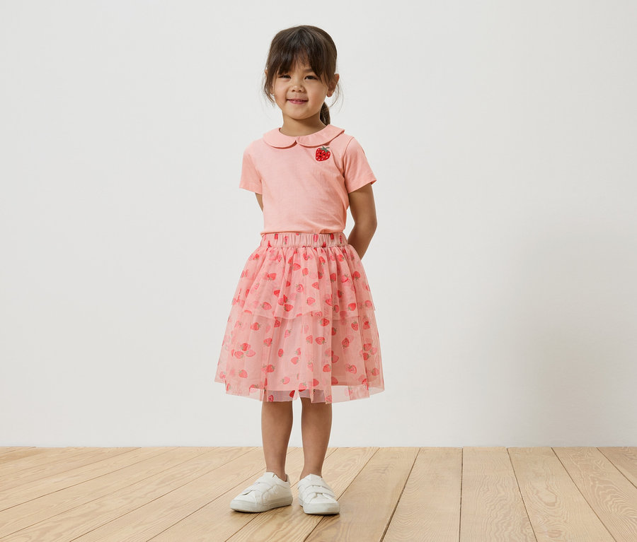 Egy kislány áll egy fehér fal előtt, rózsaszín, epermintás pólót, rózsaszín tüllszoknyát eperrel és fehér tornacipőt visel.