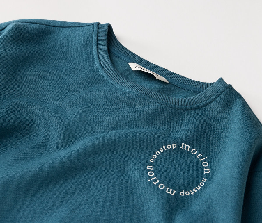 Közelkép egy petróleumkék színű pulóverről, melyen kör alakban a "nonstop motion" felirat látható.