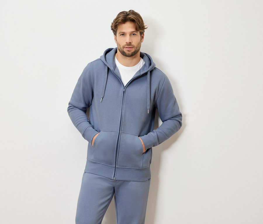 Világosbarna hajú férfi kék, kapucnis, cipzáros tréningruhában és fehér pólóban áll, mindkét keze a zsebében.