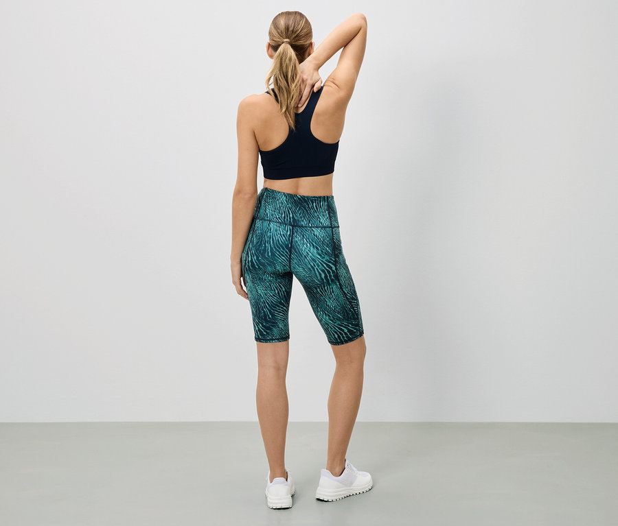 Egy nő háttal áll a kamerának, fekete sportmelltartót és rövid, mintás leggingset visel.