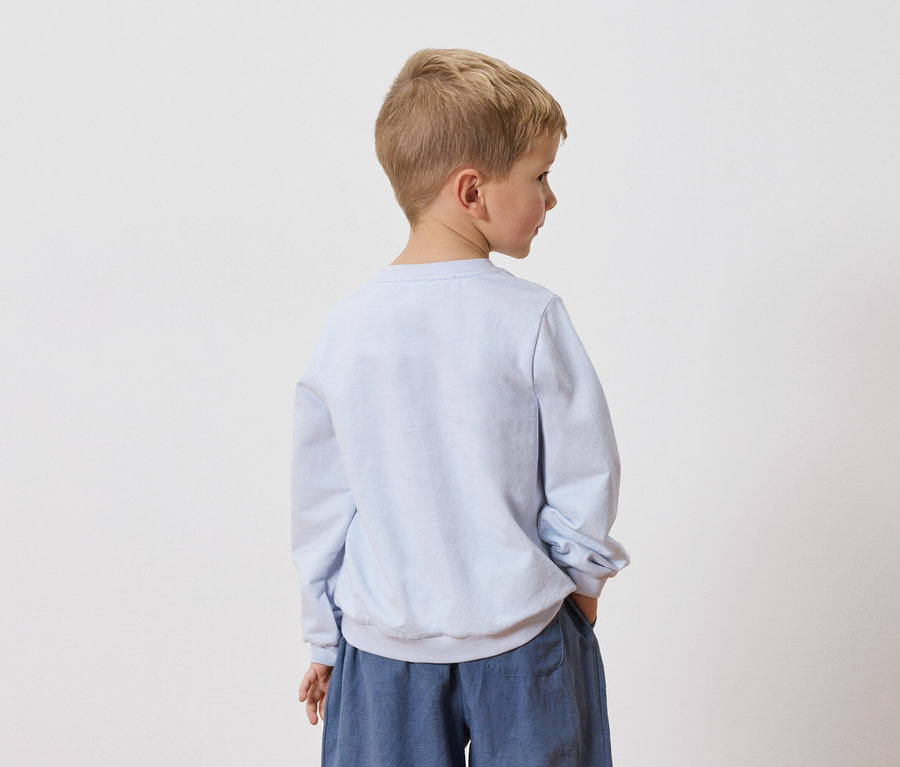 Egy szőke hajú fiú háttal áll a kamerának, világoskék pulóvert és kék nadrágot visel.