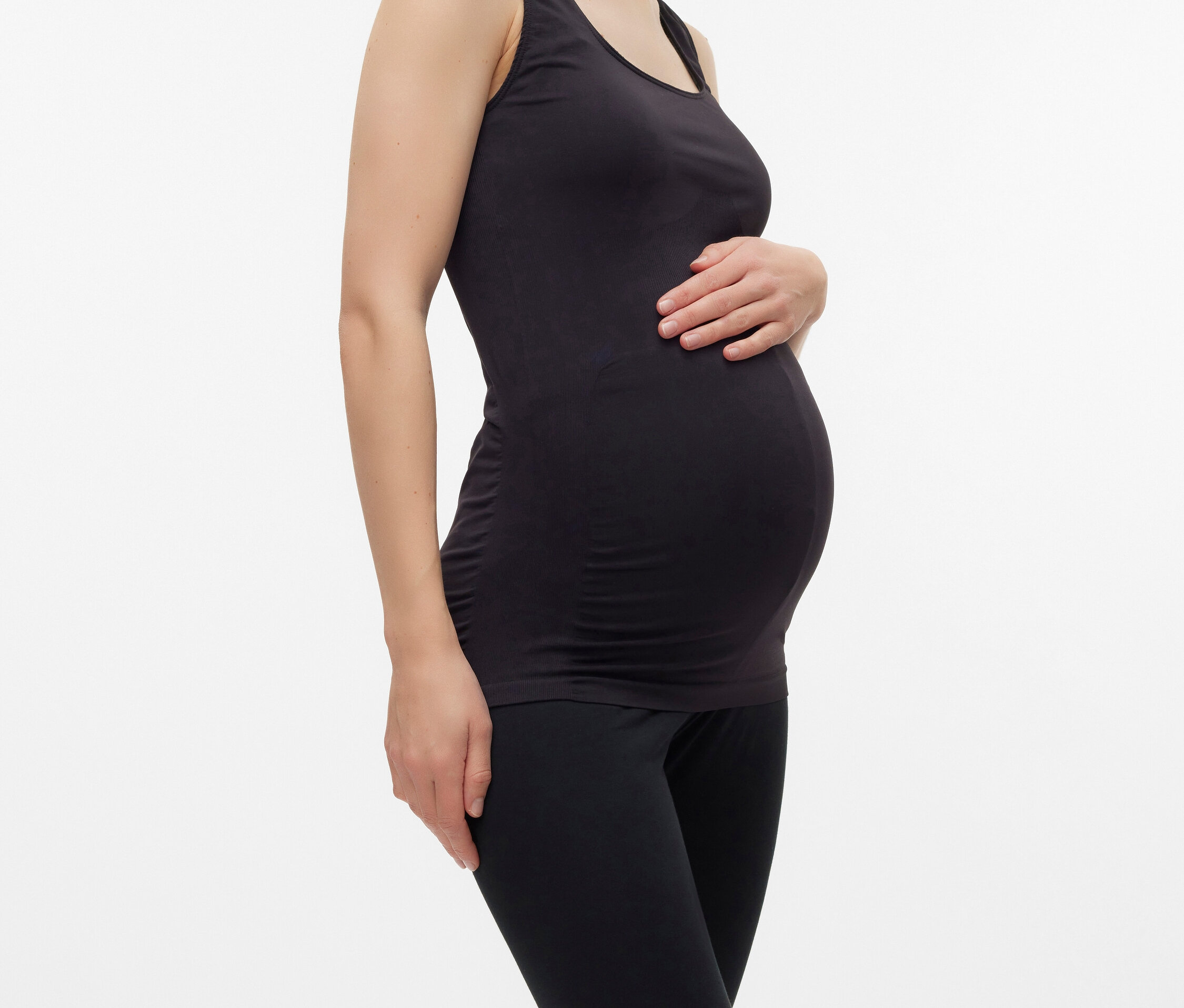 Egy terhes nő fekete trikóban és leggingsben pózol fehér háttér előtt.