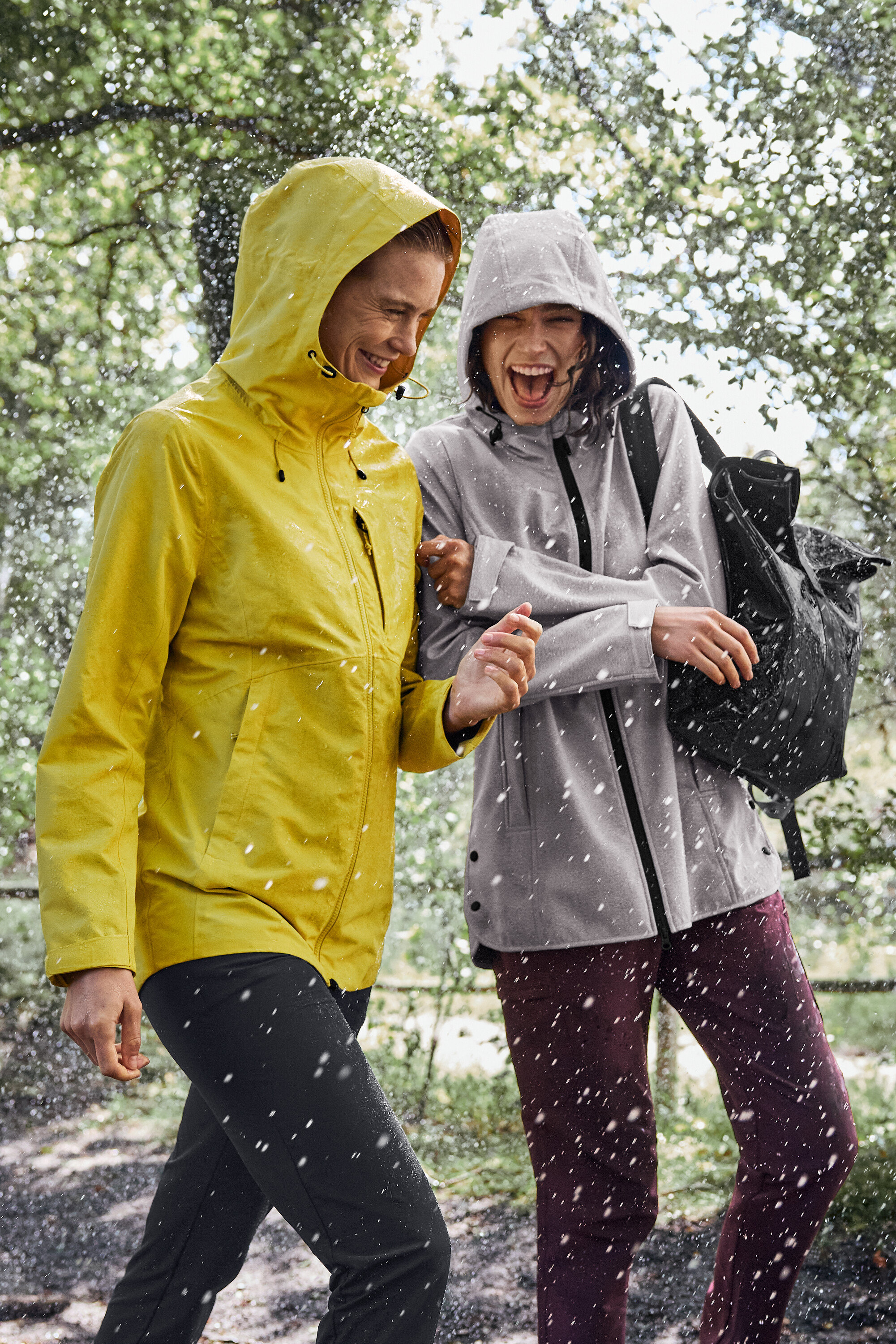 Két nő sétál az esőben, az egyik sárga esőkabátot visel, a másik pedig szürke esőkabátot fekete hátizsákkal.