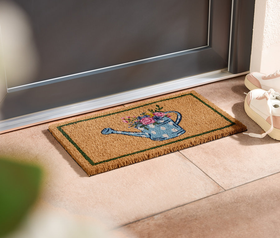 Locsolókanna mintás lábtörlő fekszik az ajtó előtt, mellette fehér tornacipők állnak.