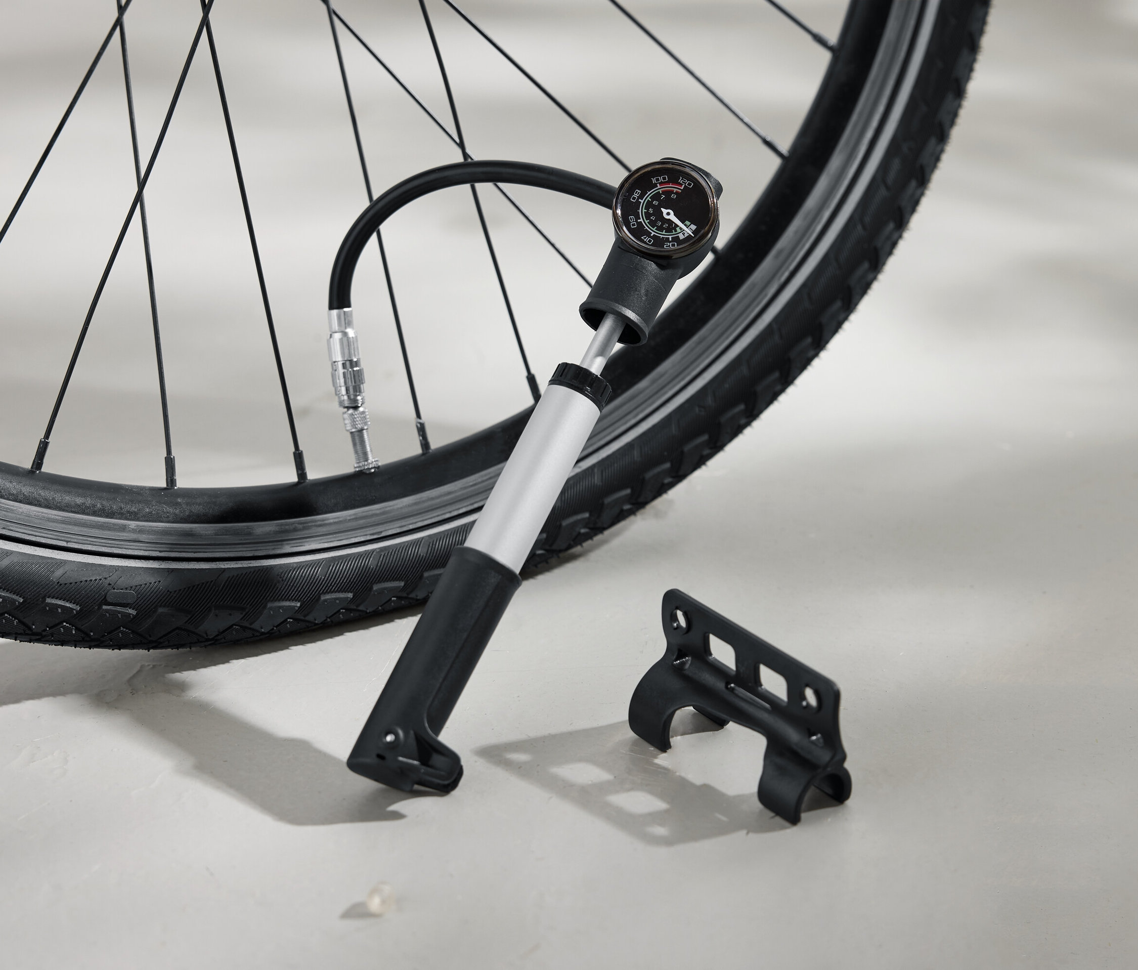 Egy kerékpár gumiabroncsát ezüst színű pumpával fújják fel, amely fekete fogantyúval és analóg nyomásmérővel rendelkezik. Mellette a földön egy fekete konzol található.