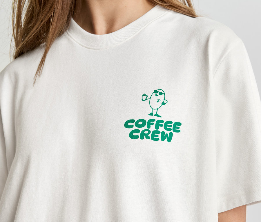 Egy világos pólót viselő személy közeli felvétele, a pólón zöld színű "Coffee Crew" felirat és egy rajzolt kávébab figura látható.