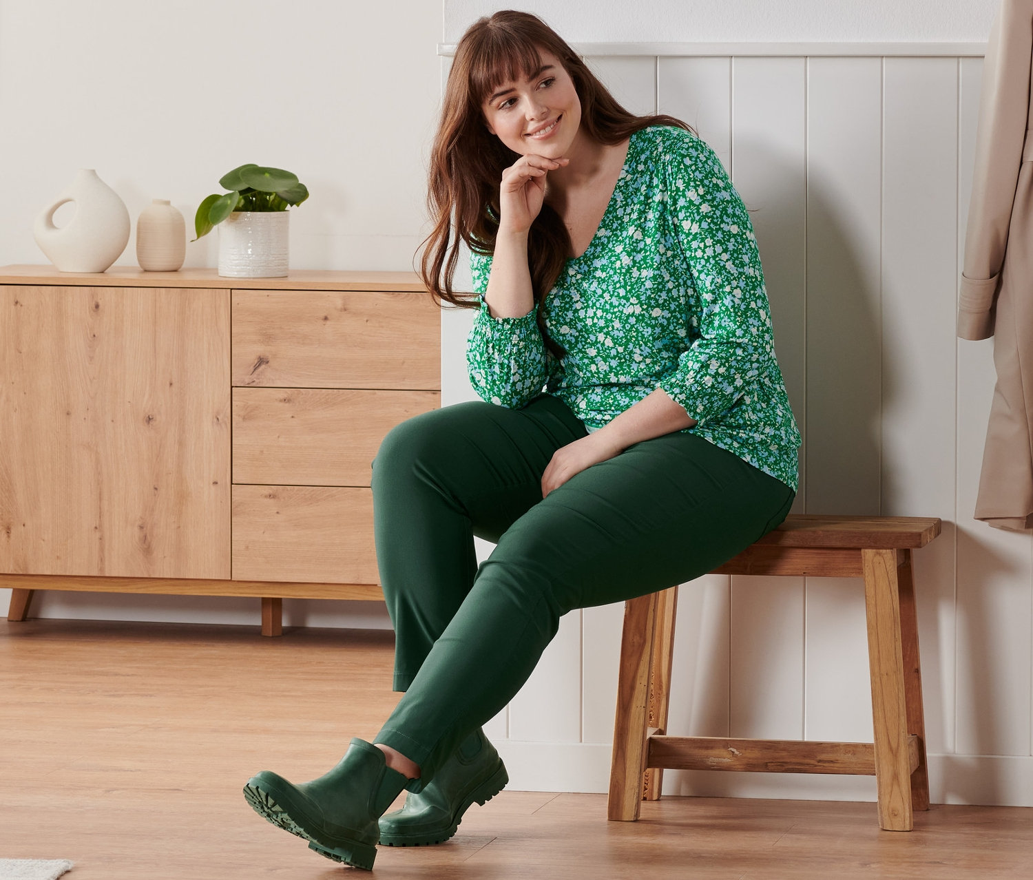 Egy nő ül egy padon és virágos, zöld, 3/4-es ujjú pólóblúzt, bengalin nadrágot és női gumi bokacsizmát visel.