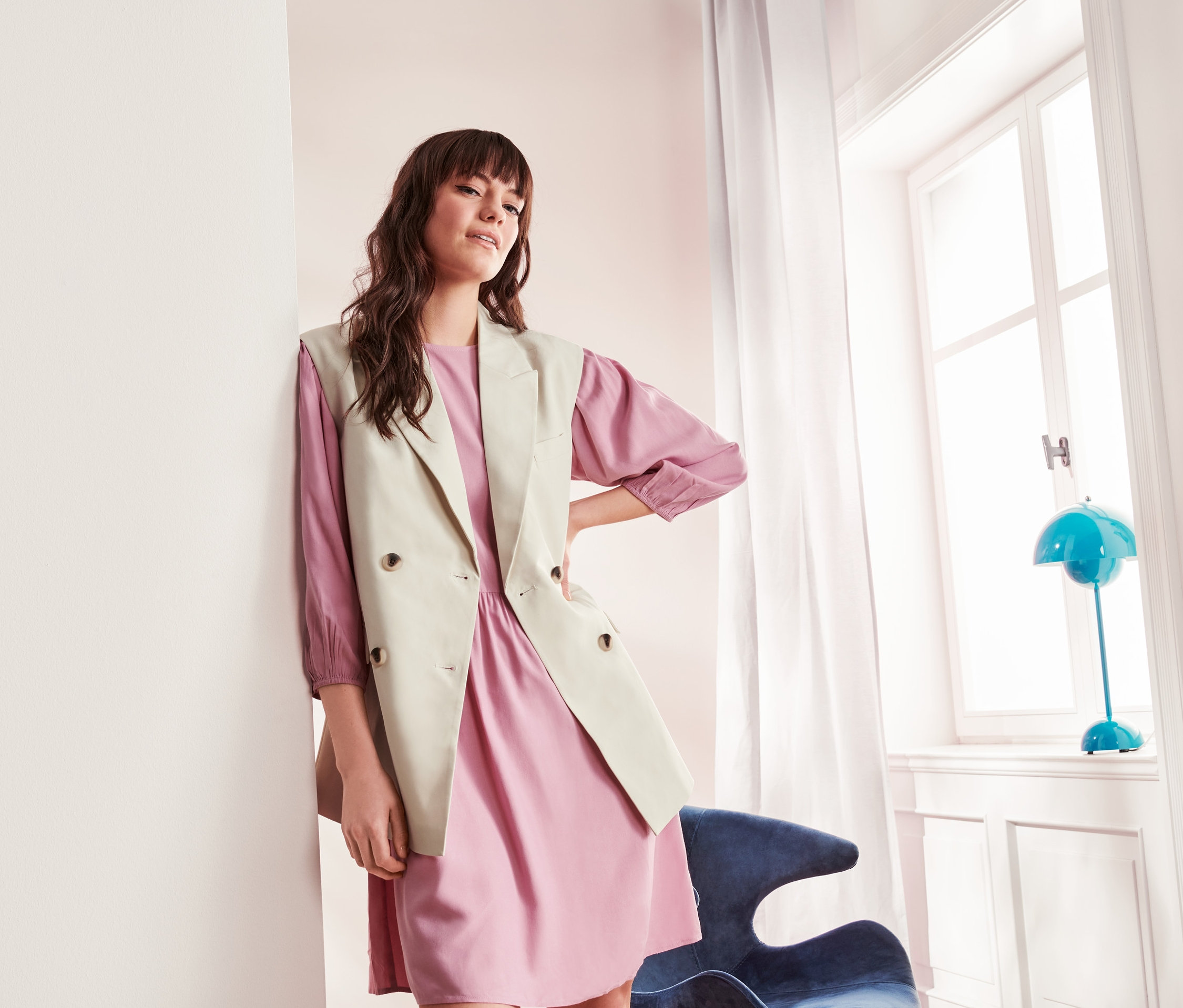 A falnak támaszkodó nő rózsaszín ruhát és női blézermellényt visel.