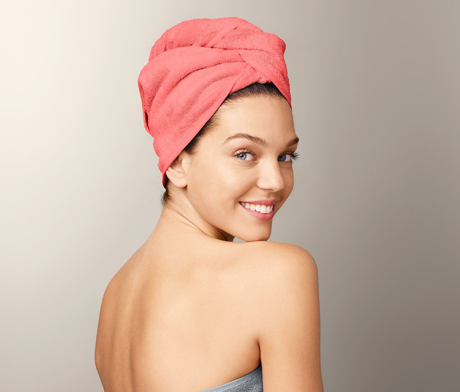 Nő mosolyog a válla fölött, törölköző turbánt visel és szürke fürdőlepedőbe van csavarva.