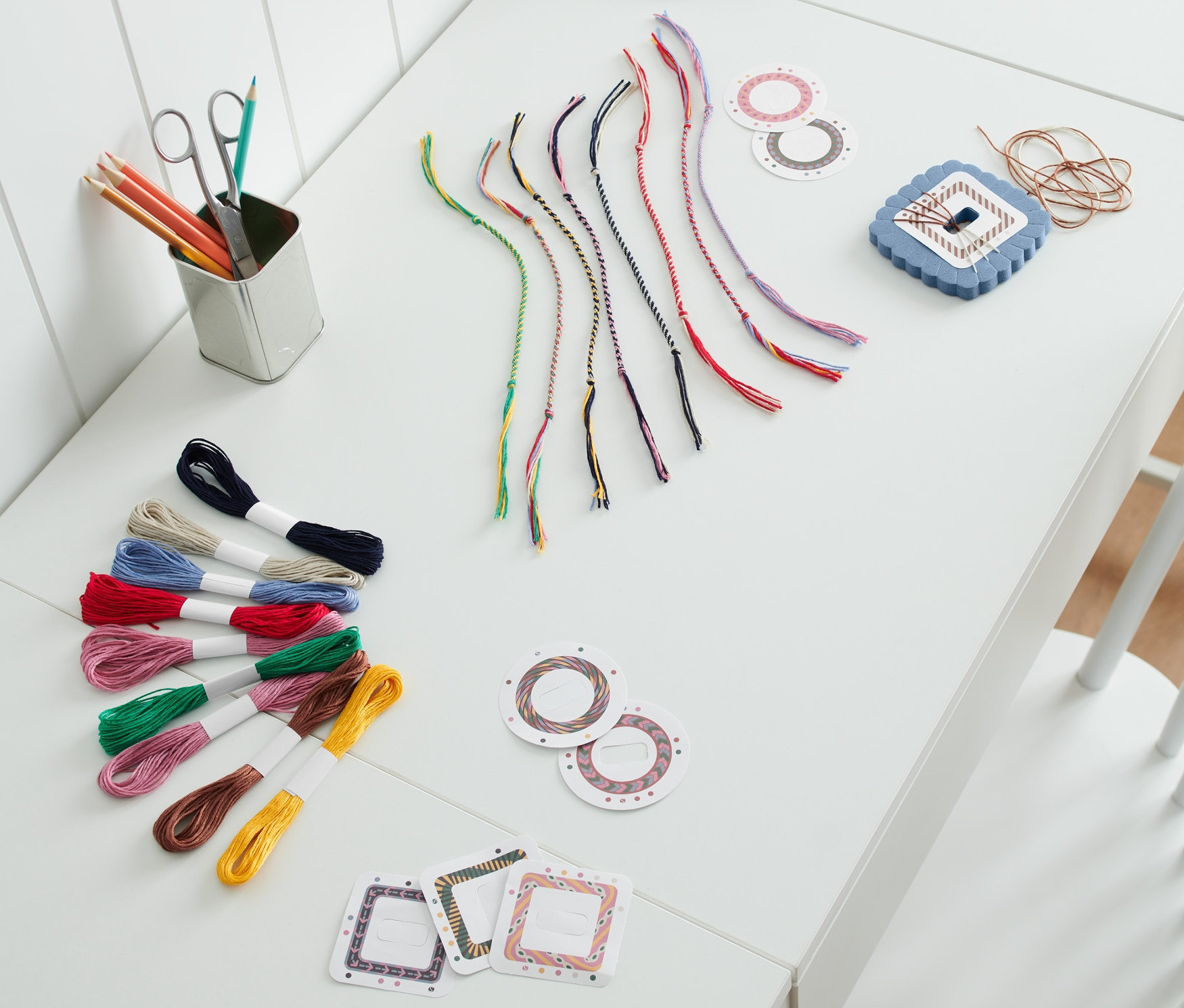 Egy fehér asztalon Barátságkarkötő készítő készlet, színes fonalak és egy kék lámpa van.