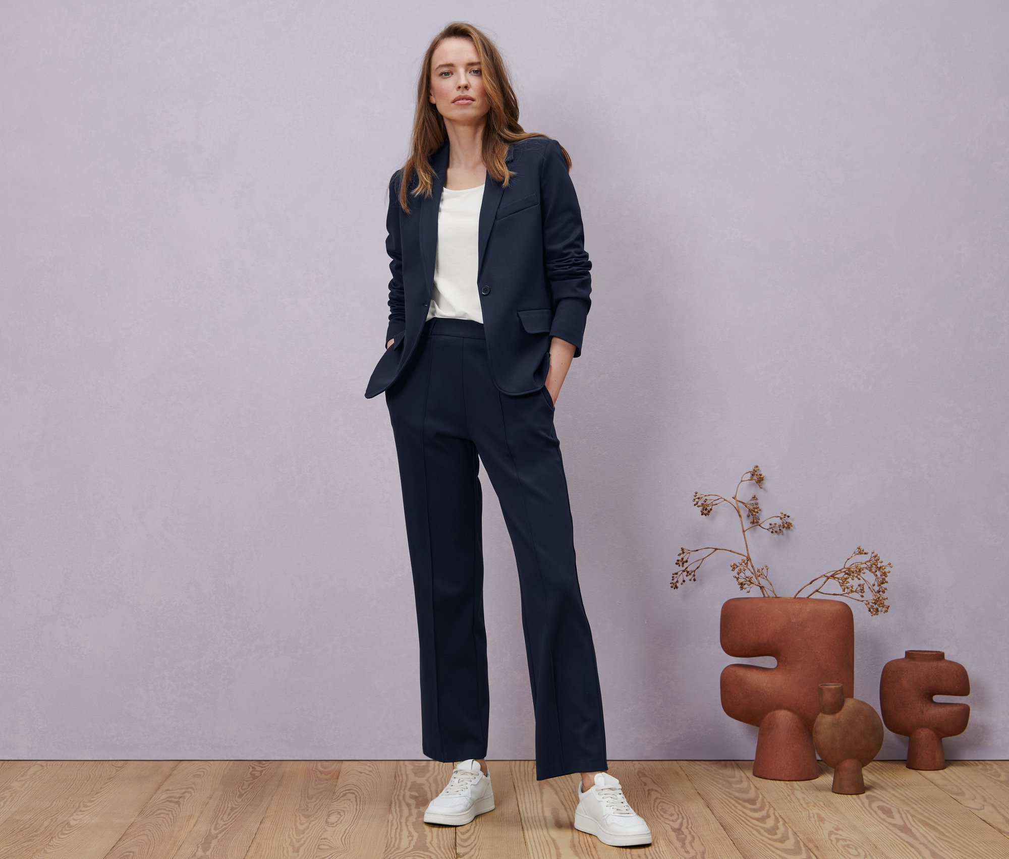 Egy nő sötétkék dzsörzénadrágban, dzsörzé blézerben és fehér, női félhosszú ujjú felsőben pózol.